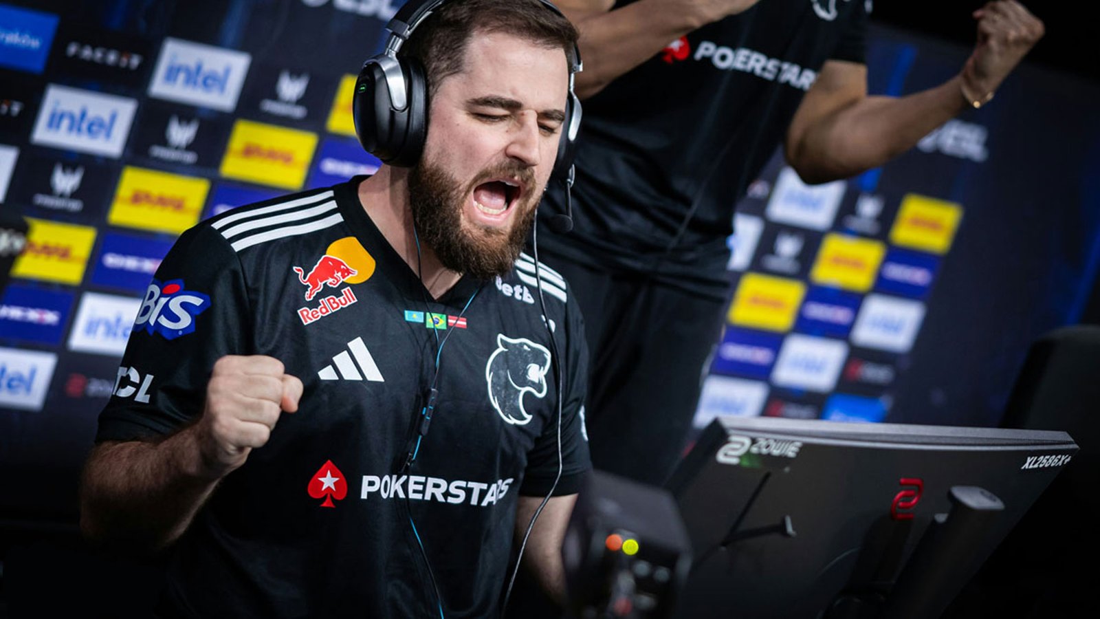 Jogador de Counter-Strike da equipe FURIA gritando e vibrando de alegria em frente ao monitor durante uma partida do IEM Krakow 2026. Ele usa uniforme preto com patrocinadores e fones de ouvido profissionais.